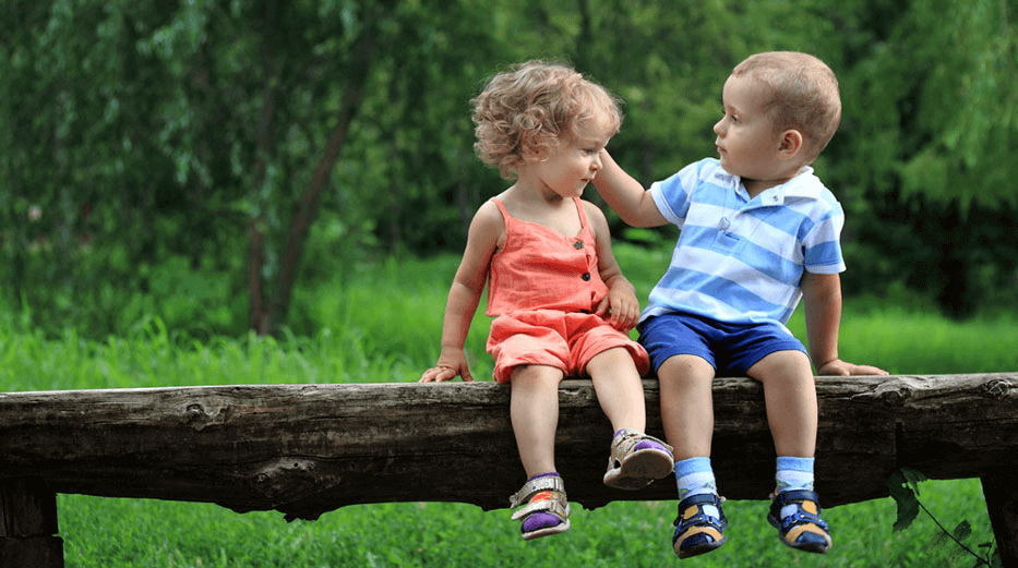 Two toddlers sit on a log bench, the boy gently brushing back the girl's curly hair with his hand.